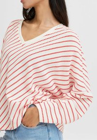 Un pull blanc avec des rayures diagonales rouges, doté d'un col en V et d'une coupe décontractée. La texture du tissu semble douce, avec des bords de manches enroulés.