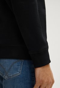 Schwarzer Sweatshirtbund mit gerippter Textur, kombiniert mit blauen Jeans, die sichtbare Nähte und ein klassisches Gesäßtaschendesign aufweisen.