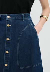 Jupe en denim bleu foncé avec taille haute, boutons en métal doré, grande poche latérale et coutures blanches contrastantes.