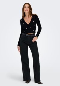 Femme portant un cardigan noir court avec des cœurs roses, un jean large noir, une ceinture noire, un collier en forme de cœur et des chaussures noires à clous, en posant.