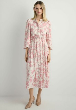 Kobieta ubrana w białą sukienkę midi w różowe kwiatowe wzory, z długimi rękawami, z przodu zapinaną na guziki, w beżowych szpilkach, stojąca na neutralnym tle.