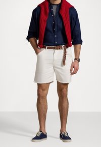 Witte denimshorts met rafelige zomen, gecombineerd met een marineblauw overhemd met knopen en een rood mouwloos vest. Bruine gevlochten riem en marineblauwe schoenen.