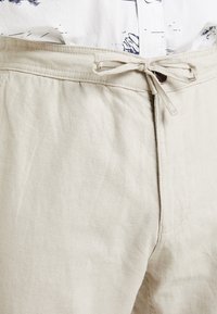 Pantalon en lin beige avec une ceinture en tissu nouée et un bouton, porté avec une chemise blanche présentant un motif abstrait bleu.