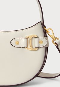 Κρεμ δερμάτινη τσάντα με καμπύλο σχήμα, διαθέτοντας χρυσές λεπτομέρειες και έναν χαρακτηριστικό κλειδαριά με δύο βρόχους. Η αντίθεση με το σκούρο περιτύλιγμα προσθέτει ορισμό.