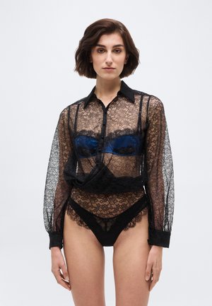 Kvinde iført en gennemsigtig sort blondebluse over en mørkeblå bh og matchende sorte blondeundertøj, stående foran en ensartet lys baggrund.