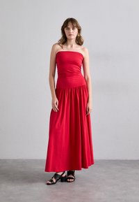 Czerwona, bezramionowa maxi sukienka wykonana z miękkiego materiału, z dopasowaną górą i zwiewnym, plisowanym dołem. Nosi się z czarnymi sandałami na słupku.