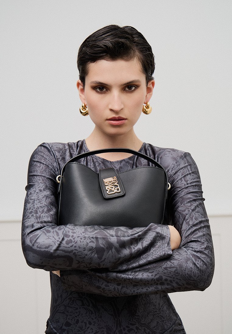 Sac à main en cuir noir avec une forme structurée et des accessoires en or. Le sac présente un fermoir proéminent et est tenu devant un mannequin portant un haut à motifs.