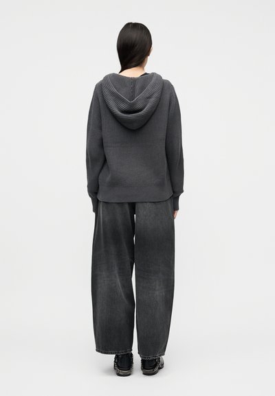 Personne vue de dos portant un sweat à capuche en maille gris foncé, un jean large noir délavé et ample, ainsi que des baskets noires avec un détail en chaîne argentée.