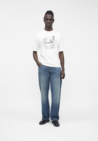 Weißes T-Shirt mit Grafikdruck, das ein Berg- und Blumenmotiv zeigt, kombiniert mit hellblauen, weit geschnittenen Jeans und schwarzen Schuhen, stehend vor einfachem Hintergrund.