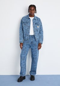 Hellblaues Jeansjackett mit einem dezenten Muster, kombiniert mit passenden hellblauen gemusterten Hosen. Schwarze Schuhe runden das Outfit ab.