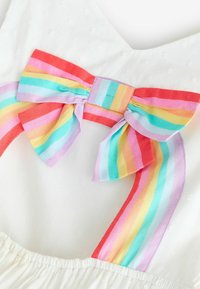 Tela blanca con sutiles puntos que presenta un gran lazo anudado en el cuello con rayas de arcoíris, mostrando franjas rojas, rosas, amarillas, verdes, azules y moradas.