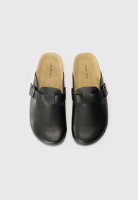 Sabots noirs avec un extérieur en cuir lisse, un bout arrondi et une sangle à boucle sur le côté. L'intérieur est doté d'un coussin doux de couleur tan.