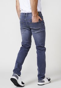 Jeans de mezclilla azul oscuro con corte ajustado. Cuenta con dos bolsillos traseros con costuras decorativas y costuras visibles. Se usan con zapatillas blancas.