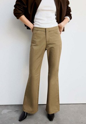 Persona con pantalones acampanados beige, top blanco, chaqueta oscura y zapatos negros puntiagudos, de pie con las manos en los bolsillos.