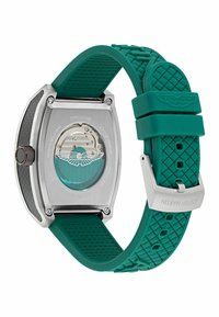 Aston Martin pulkstenis ar titāna korpusu un zaļu gumijas siksniņu. Īpašības ietver teksturētu dizainu un caurspīdīgu aizmuguri, kas atklāj mehānismu.