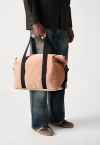 Peachfarget duffelbag laget av glatt vanntett materiale, med svarte håndtak og detaljer, med glidelås på toppen og logodesign.