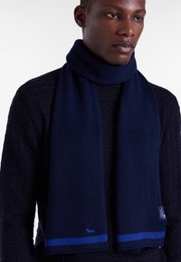 Mørkeblå uldhalstørklæde med ribbet tekstur og en kontrastfarvet blå stripe i enderne; har et lille logo i blå tråd.