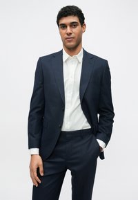 Tengerészkék öltönykabát hozzáillő nadrággal, alatta sima fehér gombos ing. Karcsúsított szabás, sima textúra és minimális díszítés.