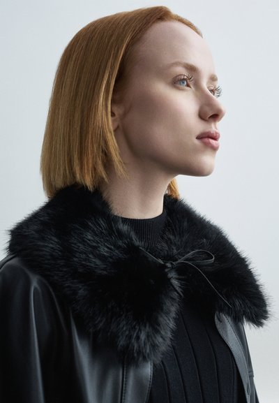 Zwarte leren jas met een pluizige zwarte nepbonten kraag en een striksluiting. Gedragen over een gestructureerde zwarte trui.