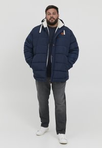 Marineblå pufferjakke med en blød hvid foring, lynlås foran og snoretræk i hætten. Sammen med grå jeans og hvide sneakers.