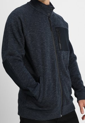 Mann in einer dunkelblauen Zip-Jacke mit schwarzen Ellbogenpatches und einer Reißverschlusstasche auf der Brust, die Hände in den Seitentaschen.