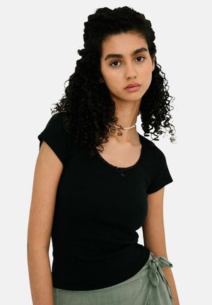 Młoda kobieta o kręconych ciemnych włosach, ubrana w czarną bluzkę z krótkim rękawem i zielone spodnie z wiązaniem w pasie, patrzy prosto przed siebie na białym tle.