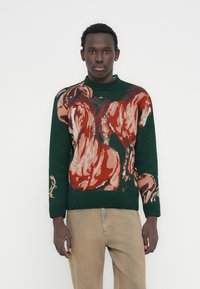 Άνδρας που φοράει σκούρο πράσινο πουλόβερ με μεγάλα κόκκινα και μπεζ σχέδια αλόγων, σε συνδυασμό με ανοιχτό καφέ παντελόνι, στέκεται μπροστά σε λευκό φόντο.