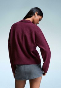 Vrouw met lang donker haar die zilveren oorbellen in de vorm van een ring draagt, een losse bordeauxrode trui en een korte grijze rok, zijwaarts kijkend tegen een effen achtergrond.