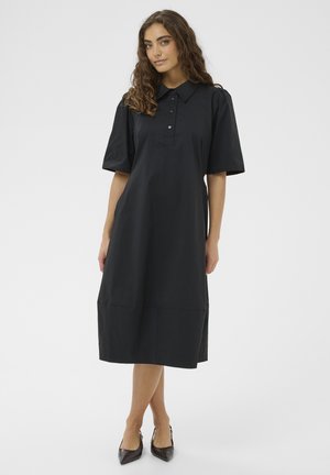 Žena s vlnitými vlasmi, oblečená v čiernych midi šatách s krátkymi naberanými rukávmi, golierom, gombíkmi a čiernymi špicatými lodičkami, stojaca na bielom pozadí.