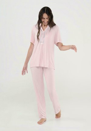 Mujer sonriendo y caminando descalza, vestida con una camiseta de manga corta color rosa claro y pantalones de pijama holgados a juego, sobre un fondo blanco.