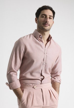 Camisa de botones de color rosa claro con rayas verticales blancas, de tela de algodón, cuello extendido y mangas remangadas. Combinada con pantalones plisados rosas.