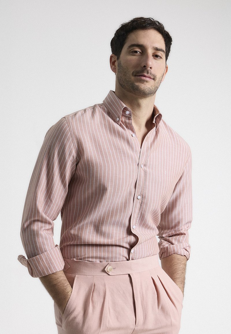 Camisa de botones de color rosa claro con rayas verticales blancas, de tela de algodón, cuello extendido y mangas remangadas. Combinada con pantalones plisados rosas.