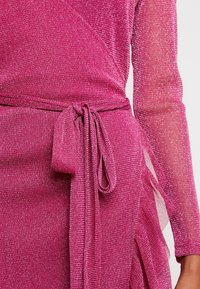 Vestido fucsia con textura brillante, mangas largas y detalle de cintura ajustada. Presenta un diseño envolvente y acentos transparentes a lo largo de los lados.
