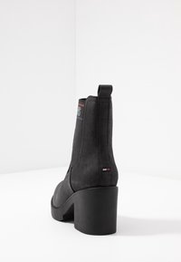 Musta mokkanahka nilkkuri korkean lohko-ullen, jossa on vetotappi ja logo-roupi punaisilla ja sinisillä yksityiskohdilla päällä.