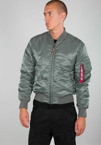 Zeleni satenski bombarder jakna s rebrastim manžetama i rubom, koja ima bočnu džepu i crvenu oznaku na rukavu.