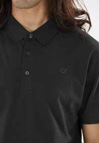 Sort polo shirt lavet af tekstureret stof, med krave og tre knapper. Subtile logo detaljer vises på venstre side.