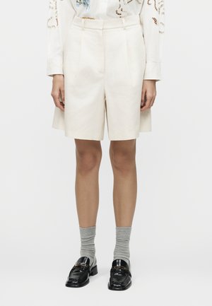 Λευκά ραμμένα σορτς μέχρι το γόνατο, συνδυασμένα με λευκό μακρυμάνικο πουκάμισο με κεντήματα, γκρι κάλτσες και μαύρα μοκασίνια με χρυσές λεπτομέρειες.