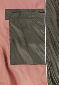 Doublure intérieure de la veste avec une grande poche rectangulaire vert olive foncé cousue sur un tissu rose, avec un rabat de fermeture velcro en haut.