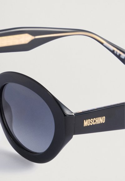 Μαύρα υπερμεγέθη γυαλιά ηλίου με στρογγυλούς φακούς, με γυαλιστερή φινίρισμα και χρυσές λεπτομέρειες. Ο λογότυπος είναι ορατός στο βραχίονα.