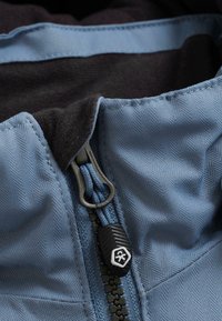 Tuvumā redzama zila jakas rāvējslēdzēja detaļa ar melnu vilkšanas cilpu, izsmeļošs šuvums un mīksts melns odere, kas redzama apkaklītes rajonā.