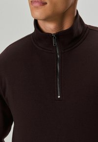 Brūns zip-up džemperis ar augstu apkakli un teksturētu audumu. Aprīkots ar metāla rāvējslēdzēju ar elegantu vilkšanas cilpu, piedāvājot vienkāršu dizainu.
