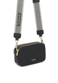 Must ristkujuline õlakott, millel on kuldsed tõmblukud ja logo, ning lai reguleeritav valge rihm mustade diagonaaltriipude ja "kazar" tekstiga.