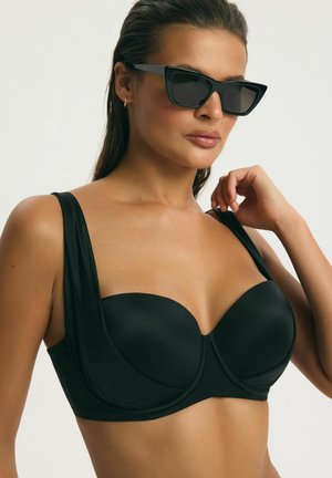 Žena v čiernom bikini s kosticou a čiernymi hranatými slnečnými okuliarmi, drží jednú ramienko pravou rukou.