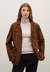 Brown suede blazer with rounded lapels, single-button closure, chest pocket, and side pockets, layered over a light turtleneck.