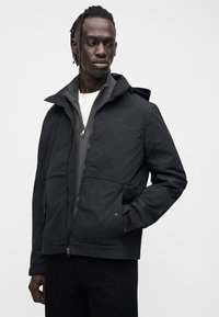 Juoda, lengva striukė su užtrauktuku, dideliu gobtuvu ir dviem priekinėmis kišenėmis. Dėvima ant pilko užtraukiamo viršutinio drabužio, derinama su tamsiomis kelnėmis.