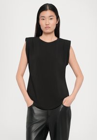 Svart ärmlös topp med strukturerade axlar, rund halsringning och mjukt tyg. Kombinerad med strömlinjeformade, glänsande svarta byxor.