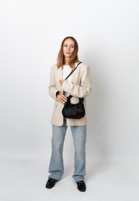 Frau trägt einen beigen Blazer, weißes Hemd, hellblaue Jeans und schwarze Schuhe, hält eine schwarze Handtasche mit Schulterriemen.