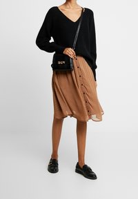 Žena v černém svetr s V-výstřihem, hnědé midi sukni na knoflíky s černými puntíky, černých mokasínech a s černou kabelkou přes rameno.