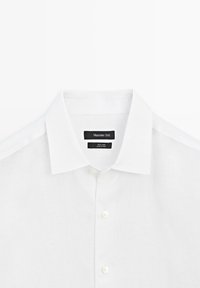 Chemise blanche en lin à boutons avec col pointu, portant des étiquettes noires indiquant "Massimo Dutti" et "100% lin, fabriqué en Chine".