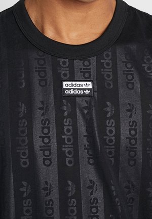 Zbliżenie czarnej koszulki Adidas z pionowym paskowanym wzorem logo i białą oraz czarną naszywką Adidas na piersi.